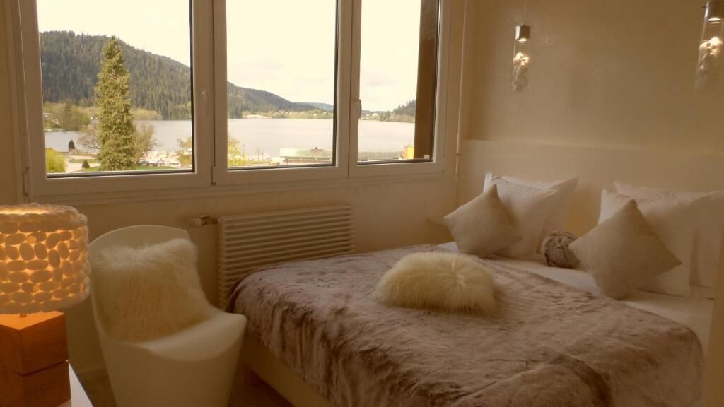 Un dormitorio con cama, una silla y vistas a un lago y montañas a través de amplios ventanales.