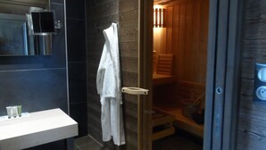 A modern bathroom with a glass-enclosed shower, a white towel hanging on the wall, and a wooden sauna visible through the glass. - hotel les loges du parc (Gérardmer)