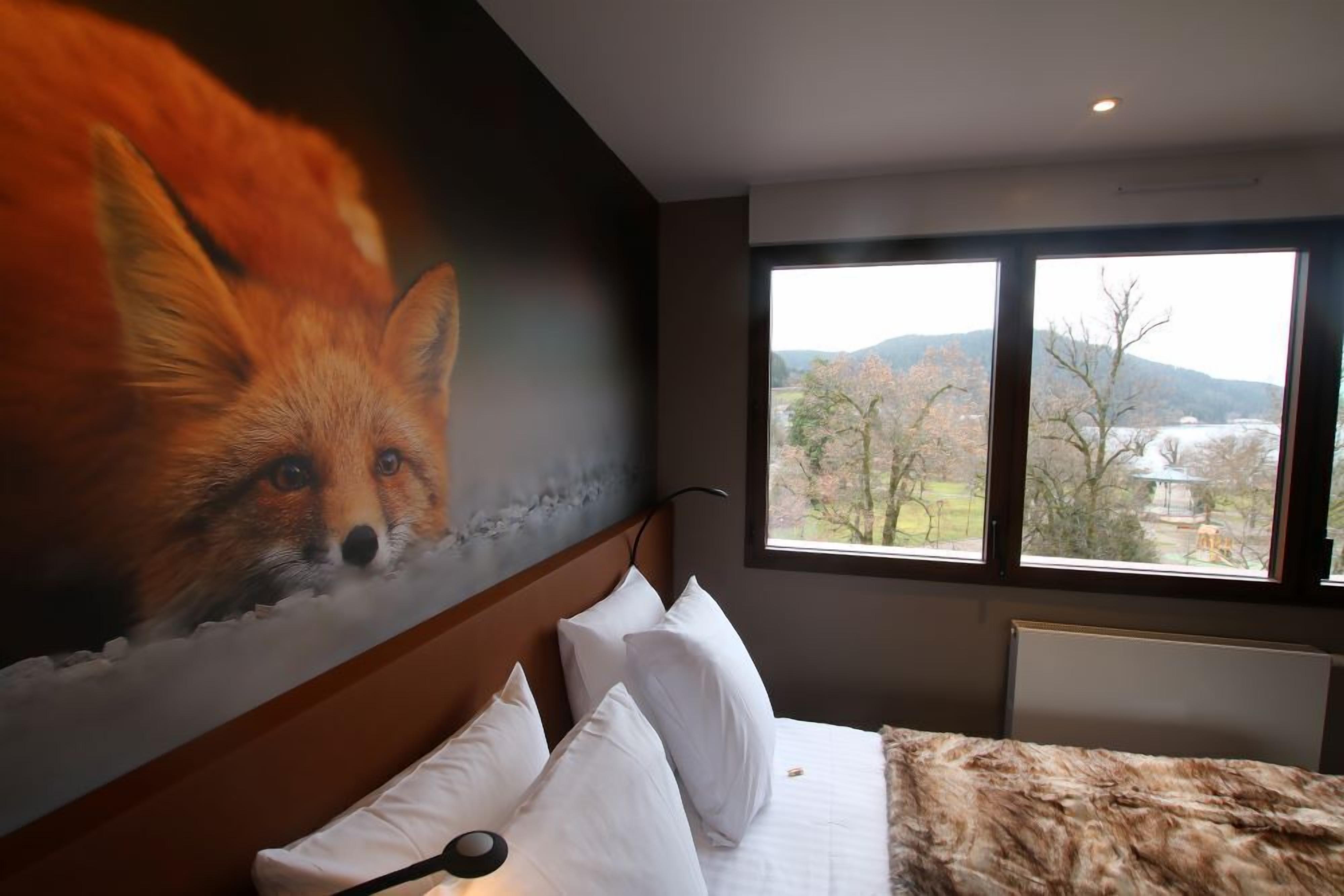 A hotel room with a large window, a bed with white bedding, and a mural of a fox on the wall.