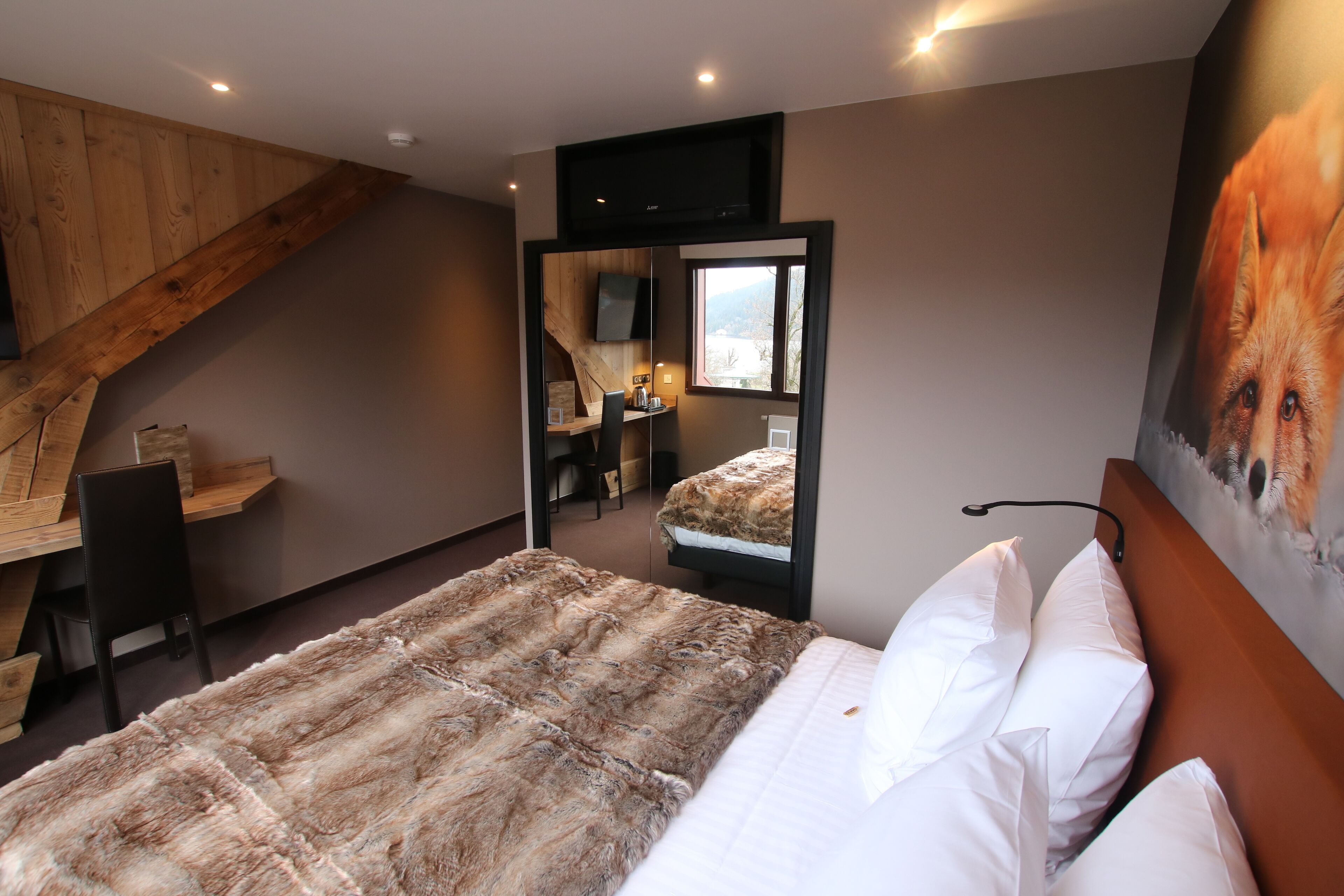 Une chambre avec un grand lit, un bureau en bois, une chaise, un téléviseur fixé au mur et un tableau représentant un renard.