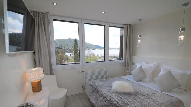A bedroom with a large window, a bed with white bedding, a bedside table with a lamp, and a view of a lake and mountains. - hotel les loges du parc (Gérardmer)