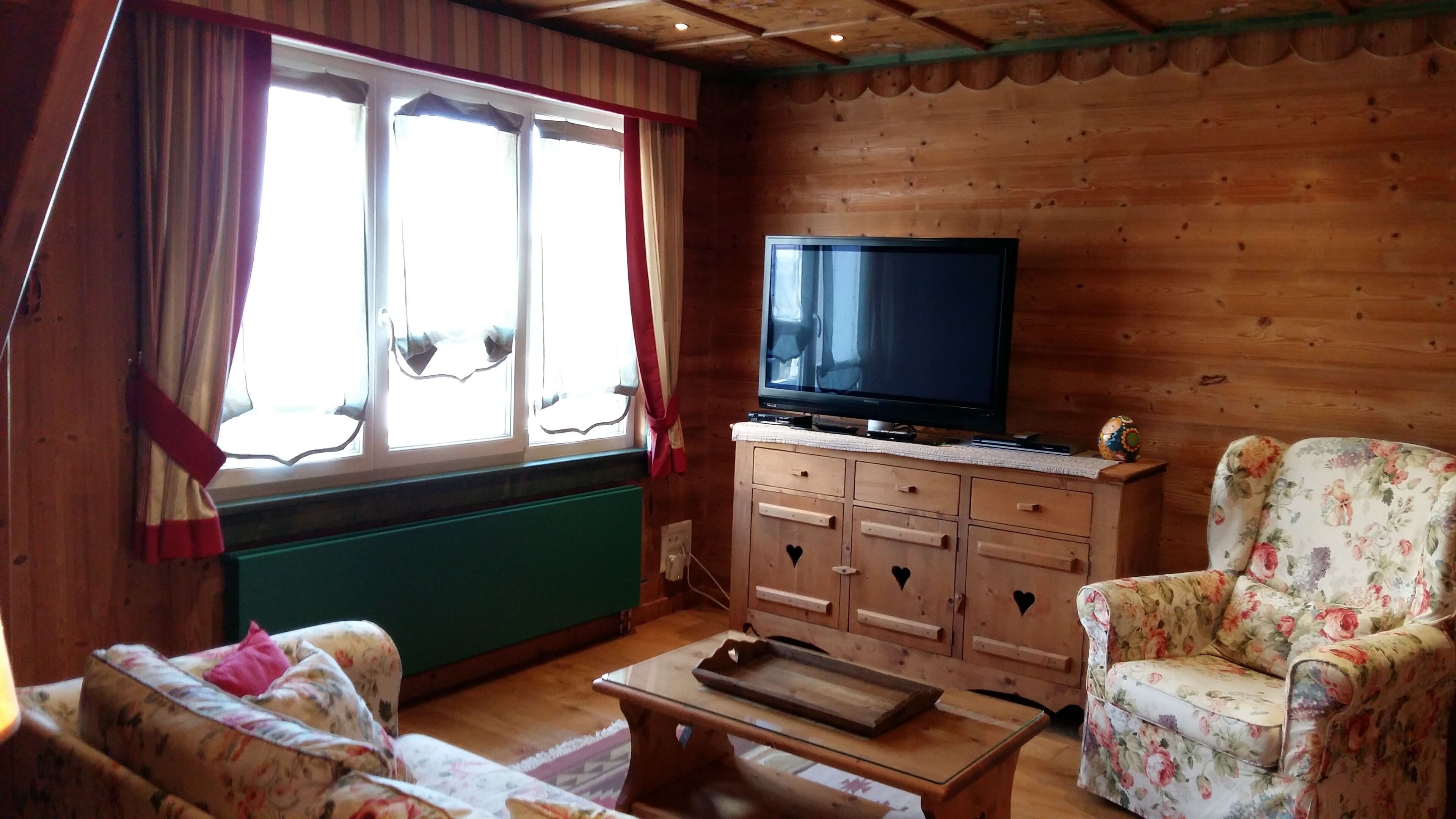 A cozy room with a wooden cabin style, featuring a television on a cabinet, floral-patterned chairs, and a window with curtains.