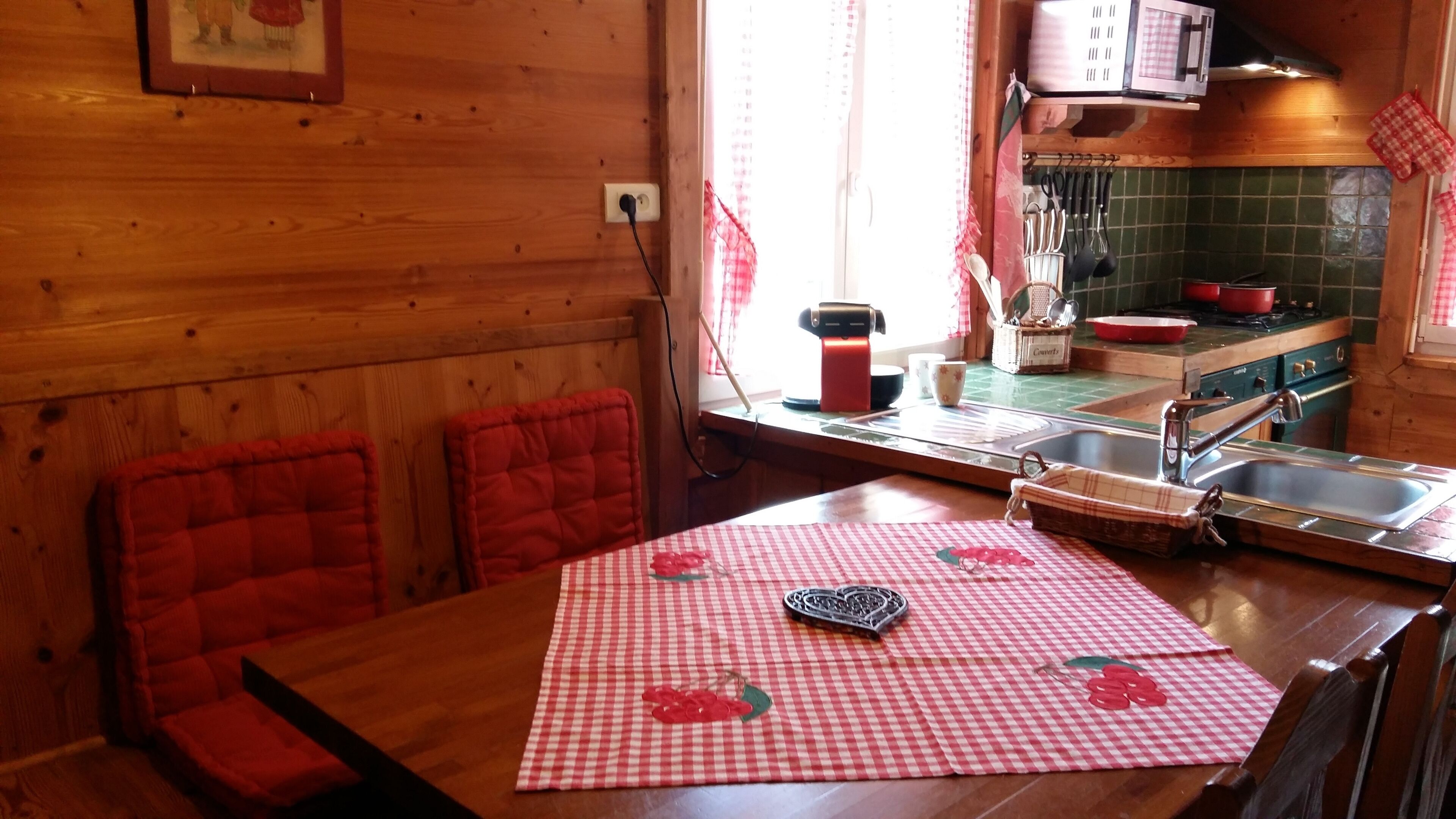A wooden interior with a dining table covered by a checkered tablecloth, red cushioned chairs, a kitchen area with a stove, sink, and various utensils.