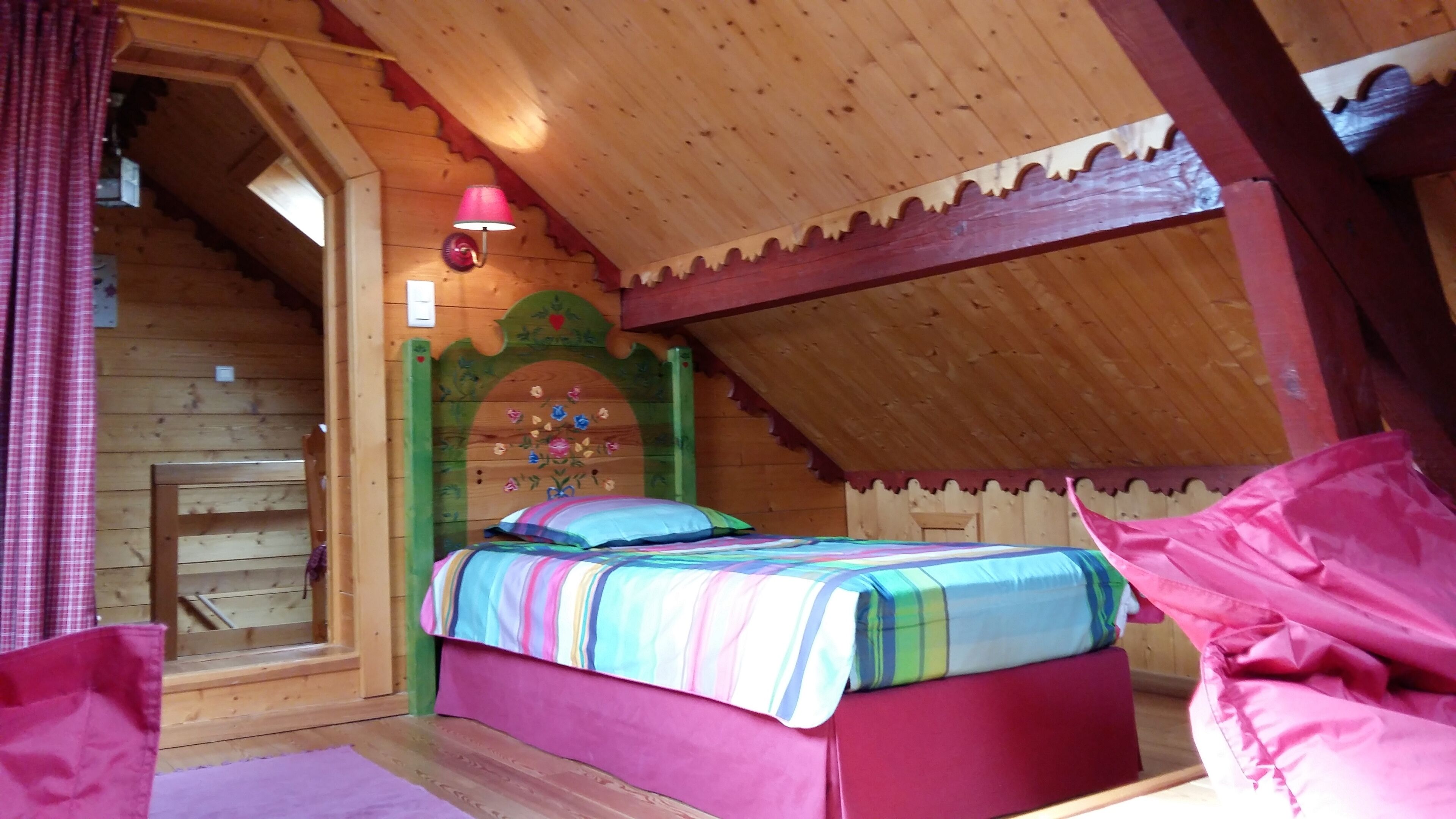 A cozy wooden cabin bedroom with a bed, a wooden headboard with floral carvings, a small wooden ladder, and a pink lamp.