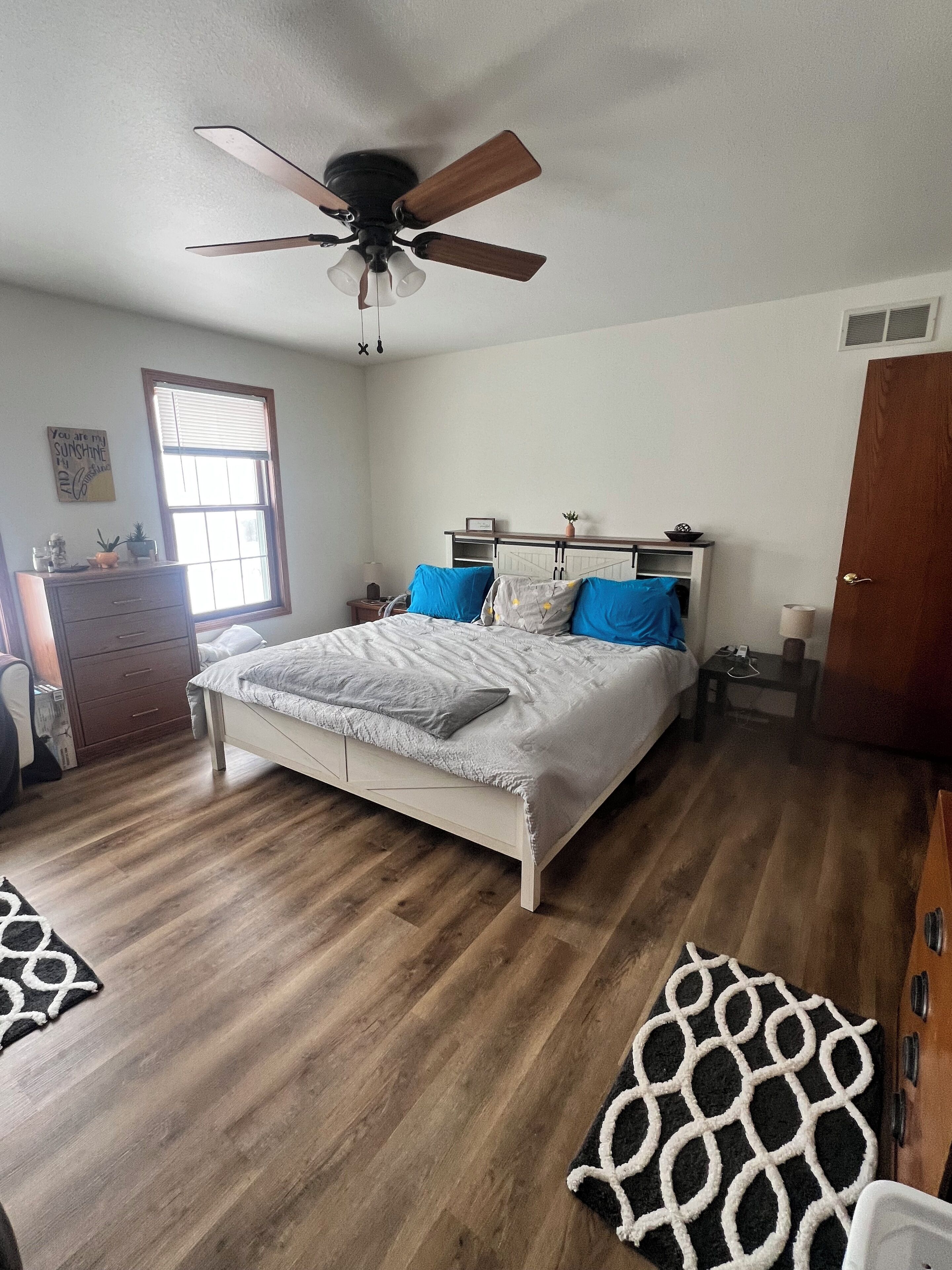 A bedroom with a bed, a ceiling fan, a window, a dresser, and a door.