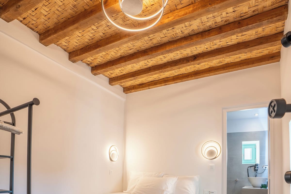 Un dormitorio con techo de madera, una cama con ropa blanca, una mesita de noche y un taburete pequeño.