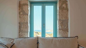 Une pièce avec un canapé beige, une table ronde sur laquelle se trouve un saladier, et une fenêtre donnant sur un paysage.