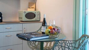 Une cuisine moderne avec des meubles blancs, un micro-ondes vert, une table à manger sur laquelle se trouvent un livre, une bouteille et un panier de fruits.