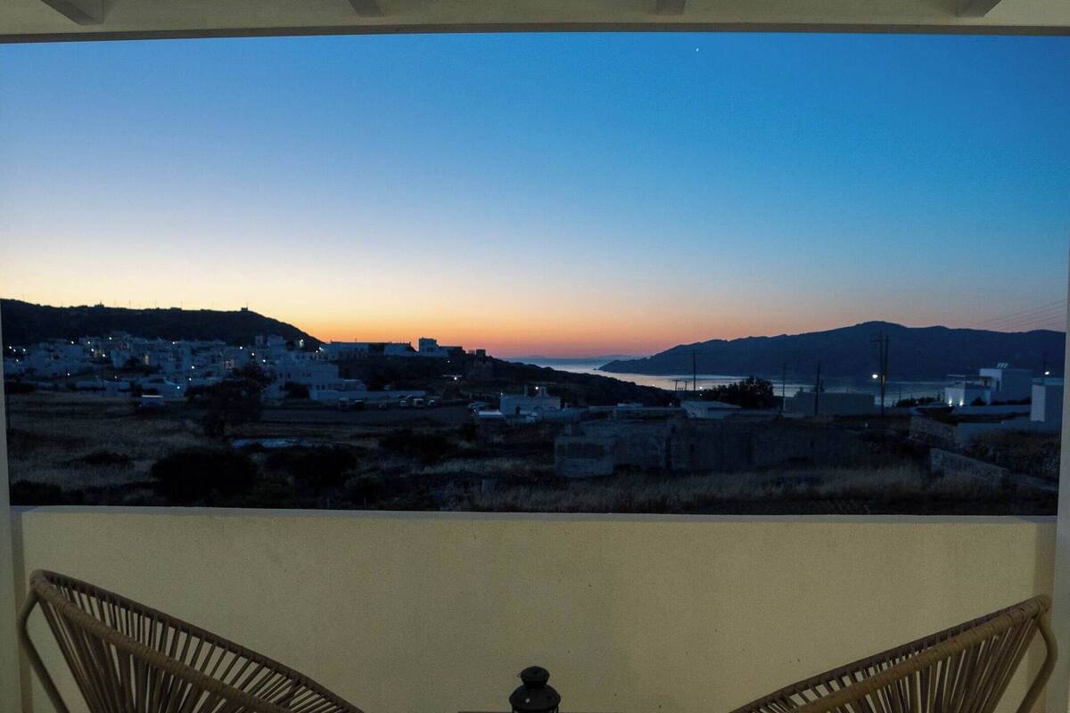 Un balcón con dos sillones de mimbre, una mesita y vista a un pueblo costero al anochecer.
