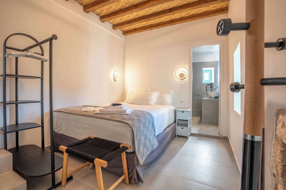 Un dormitorio con cama, mesita de noche y un pequeño banco. Hay un armario con espejo y un perchero. El cuarto tiene techo de madera y una puerta que da a un baño.