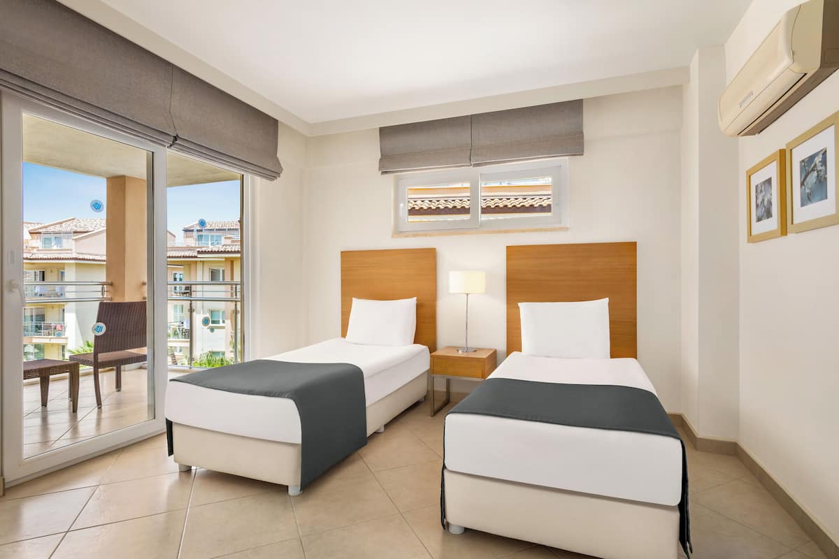 Habitación de hotel con dos camas, una ventana con vistas a edificios, una lámpara y una puerta corredera a un área exterior.