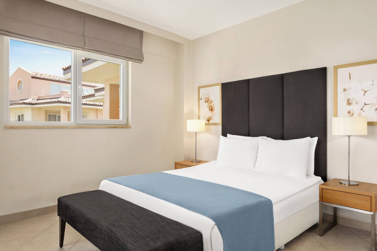 Habitación de hotel con una cama grande, un banco, dos mesitas de noche con lámparas y una ventana con vista a un edificio.