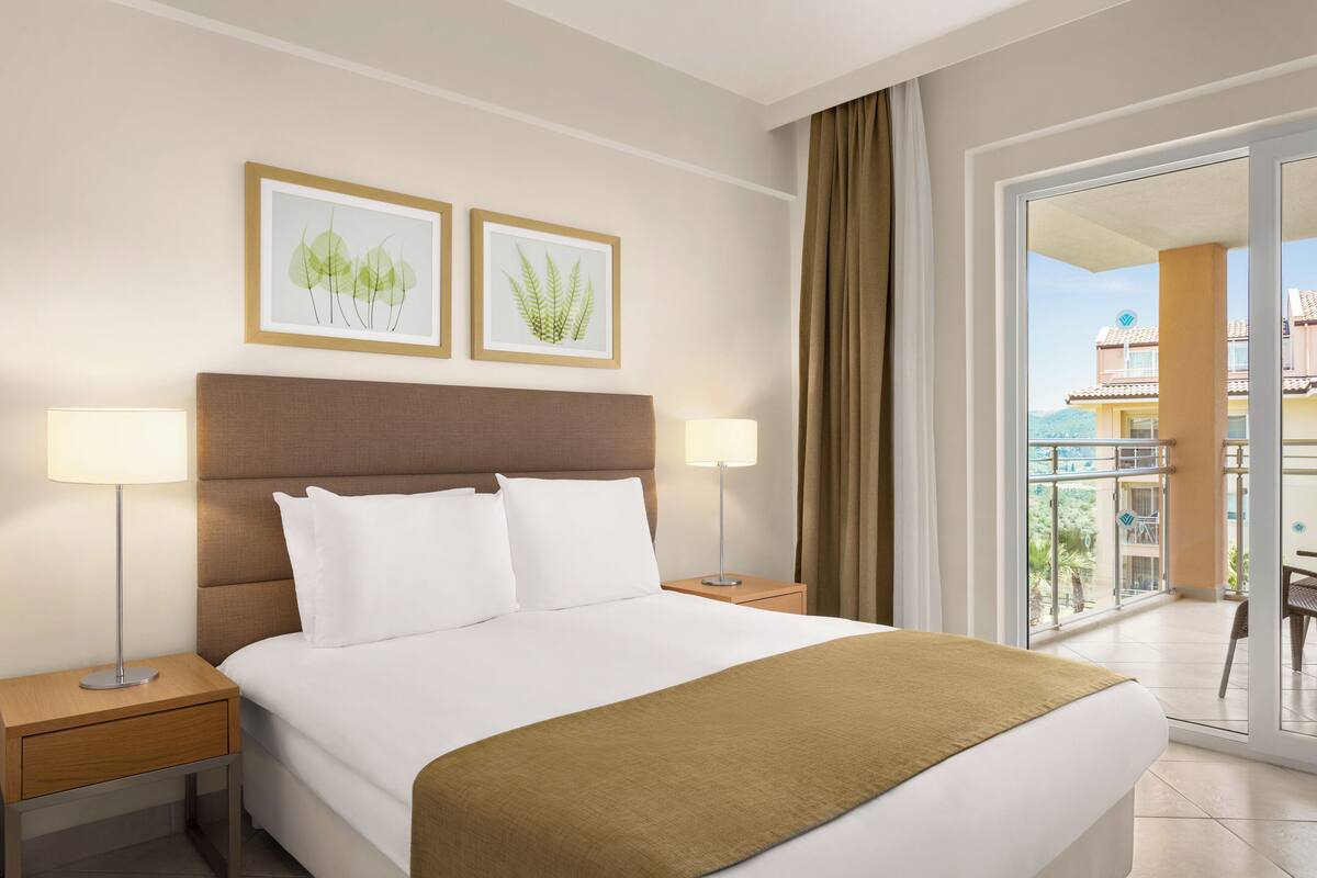 Habitación de hotel con una cama grande, dos mesitas de noche con lámparas, un cabecero marrón e ilustraciones botánicas enmarcadas en la pared.