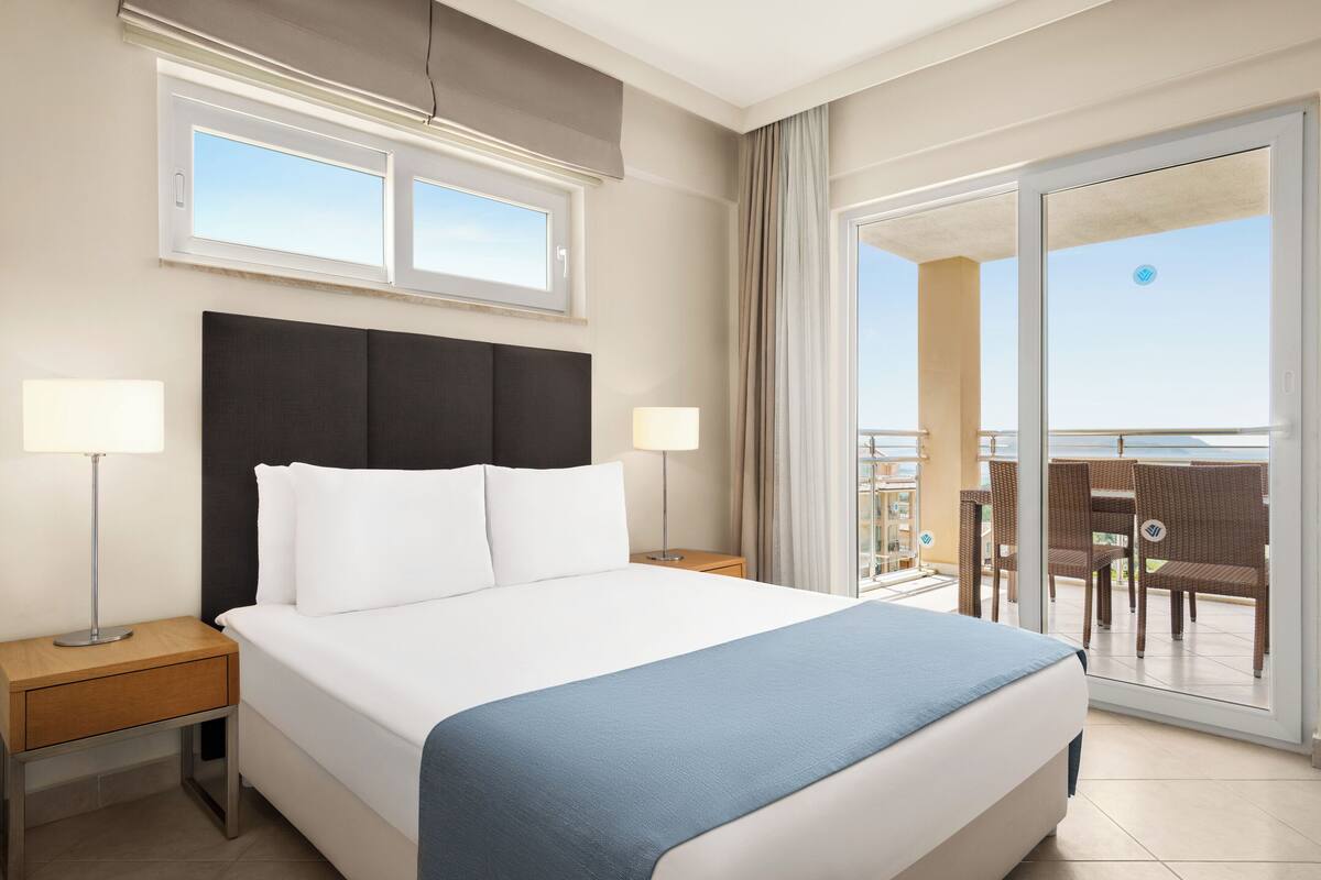 Habitación de hotel con una cama grande, dos mesitas de noche con lámparas, una ventana y un balcón con sillas.
