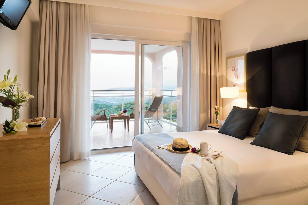Habitación de hotel con una cama grande, un escritorio con un florero, un balcón con mesa y sillas, y un televisor montado en la pared.