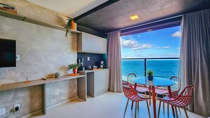 A modern kitchen with a large window overlooking the sea, a dining area with red chairs, and a flat-screen TV mounted on the wall. - Art Studio by VLV Stays (Salvador)