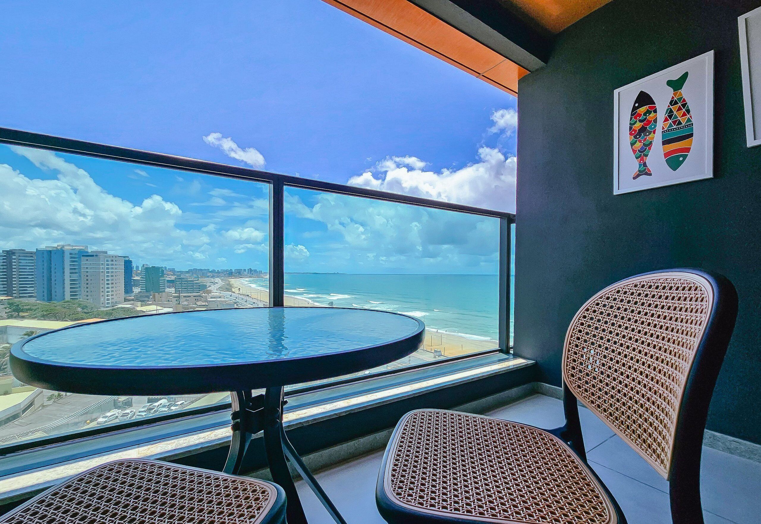 A balcony with a round table and two wicker chairs, overlooking a beach and cityscape.