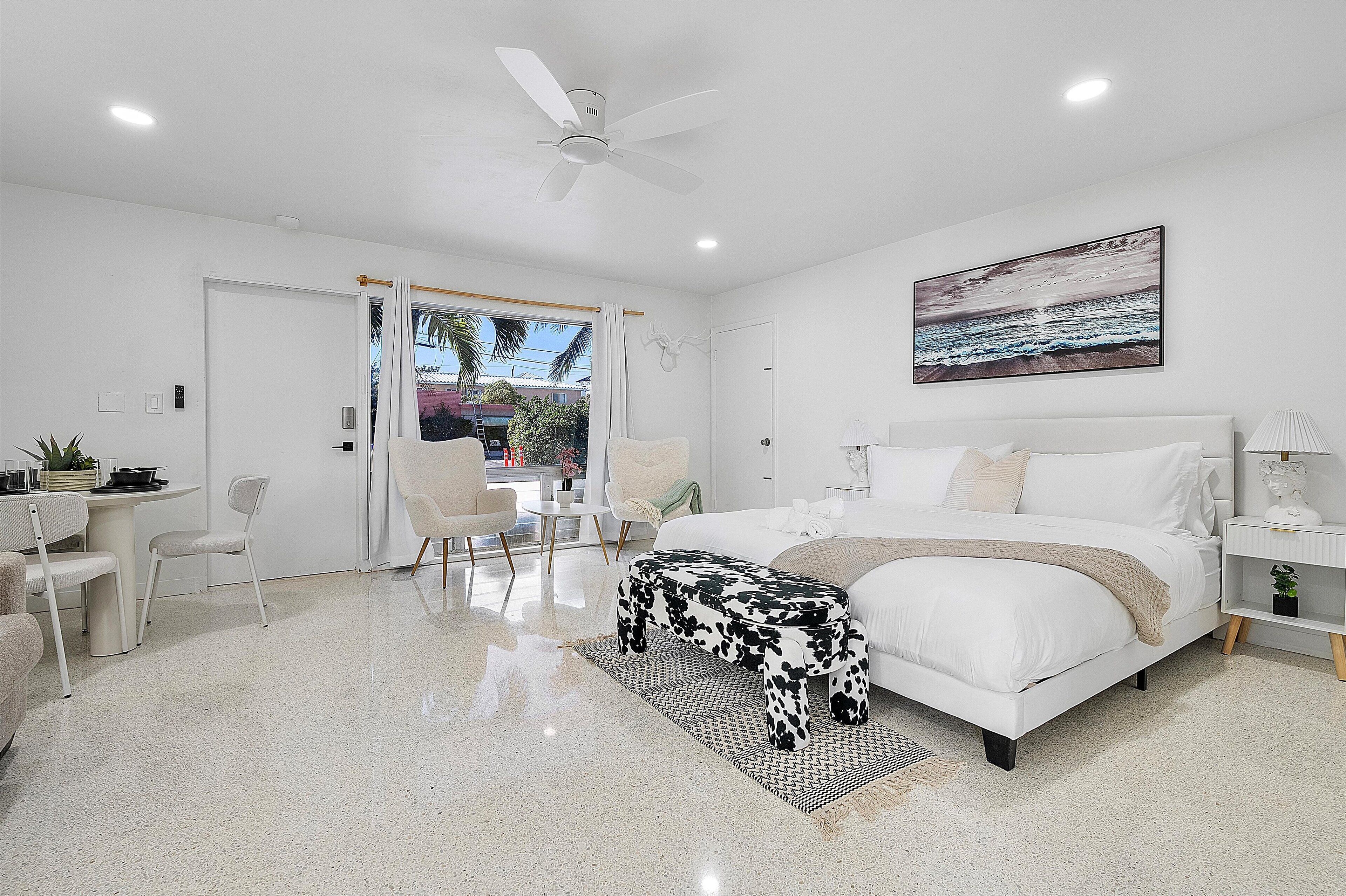 A modern bedroom with a bed, a desk, chairs, and a ceiling fan.