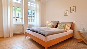 Une chambre à coucher avec un lit en bois, des rideaux blancs, un radiateur et deux tableaux encadrés accrochés au mur.