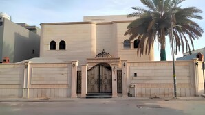 Un grand bâtiment à la façade symétrique, une entrée centrale agrémentée d’un portail décoratif et un palmier situé sur la droite.