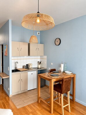 A compact kitchen with a dining area, featuring a wooden table, chairs, a hanging light fixture, and a clock on the wall. - La Cabine 17 - Studio de Standing en Centre-Ville (Boulogne-sur-Mer)
