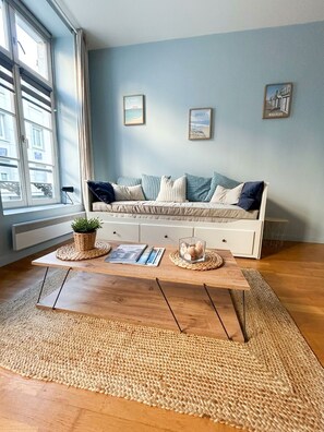 A room with a wooden coffee table, a sofa with cushions, a window with curtains, and wall art. - La Cabine 17 - Studio de Standing en Centre-Ville (Boulogne-sur-Mer)