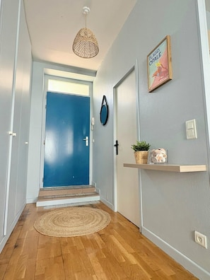 A hallway with a blue door, a hanging light fixture, a wall-mounted phone, a shelf with a plant and seashells, and a textured rug.