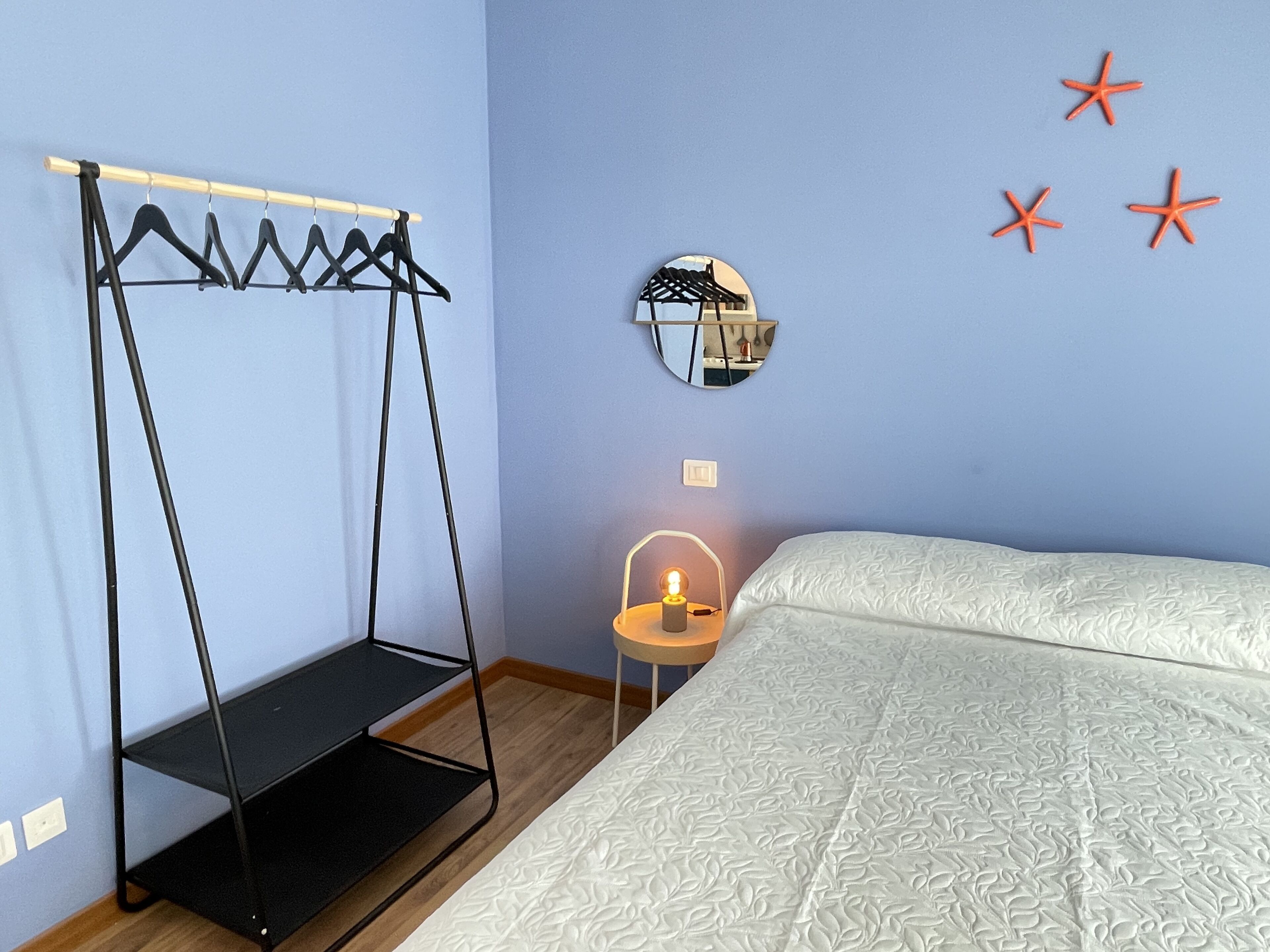 Ein Schlafzimmer mit einem Bett, einem Kleiderständer, einem kleinen Tisch mit Lampe und einem wandmontierten Spiegel.