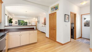 A modern kitchen with wooden cabinets, a built-in oven, and a large island. There is a dining area with a table and chairs, and a hallway leading to a door. - Rigden Retreat | Garage, Dog Friendly, Near Trails (Fort Collins)