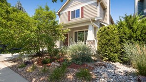 A two-story house with a front porch, surrounded by a landscaped garden with various plants and a stone pathway. - Rigden Retreat | Garage, Dog Friendly, Near Trails (Fort Collins)