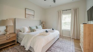 Un dormitorio con una cama grande, un ventilador de techo, una mesita de noche con lámpara, un aparador de madera y una ventana con cortinas.