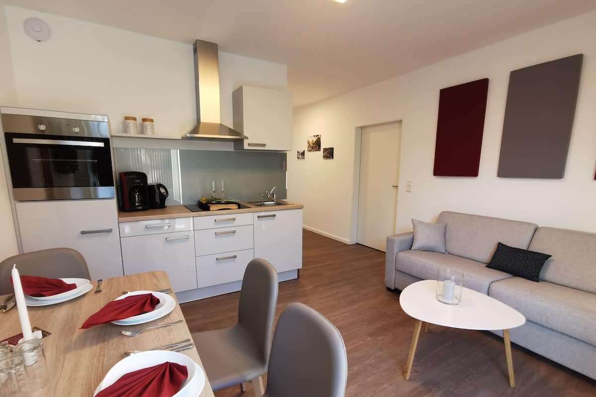 Une cuisine moderne avec des meubles blancs, un four en acier inoxydable et une machine à café. On y trouve un coin repas avec une table en bois pour quatre personnes, des chaises grises et un canapé gris avec une table basse blanche.