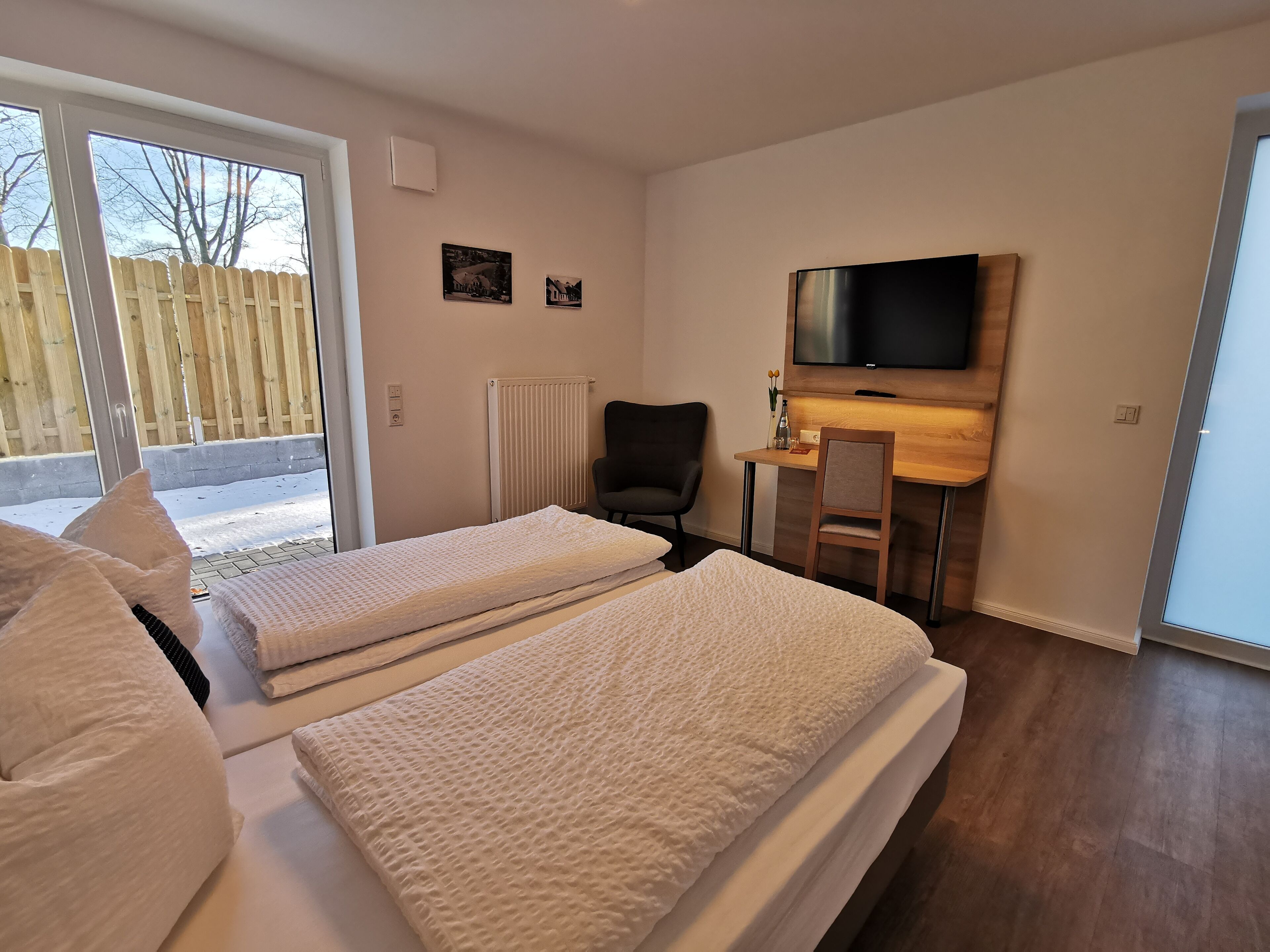 Ein Hotelzimmer mit zwei Betten, einem Schreibtisch, einem Stuhl, einem Fernseher und einem Fenster mit Blick auf einen Zaun und Bäume.