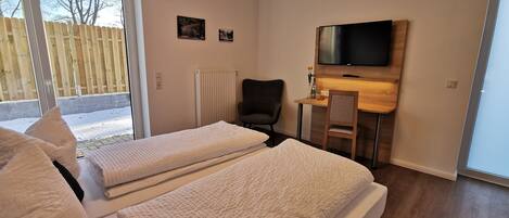 Ein Hotelzimmer mit zwei Betten, einem Schreibtisch, einem Stuhl, einem Fernseher und einem Fenster mit Blick auf einen Zaun und Bäume.