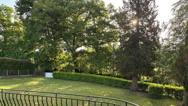 A park with a curved metal bench, well-maintained grass, and dense trees. - Family home (Eymoutiers)