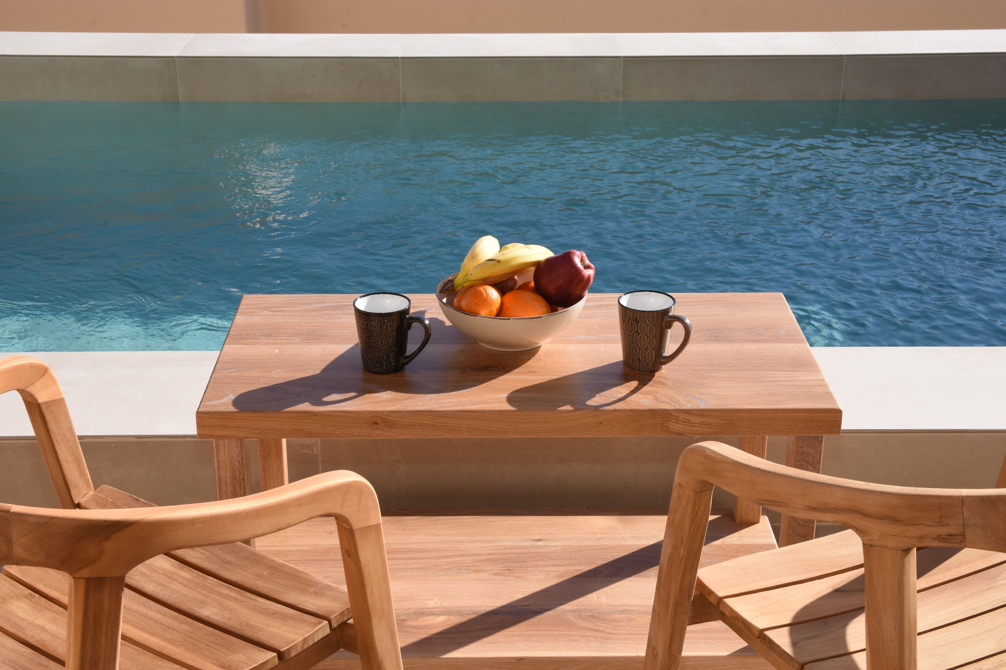 Ein Holztisch mit Obstschale und zwei gemusterten Tassen, mit Blick auf einen Swimmingpool.