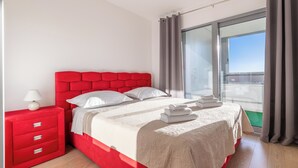 A modern bedroom with a red headboard, a white bedspread, and a view of the sky through a large window.
