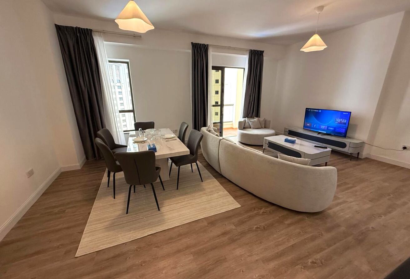 A modern living room with a dining area, a sofa, a flat-screen TV, and a large window with curtains.