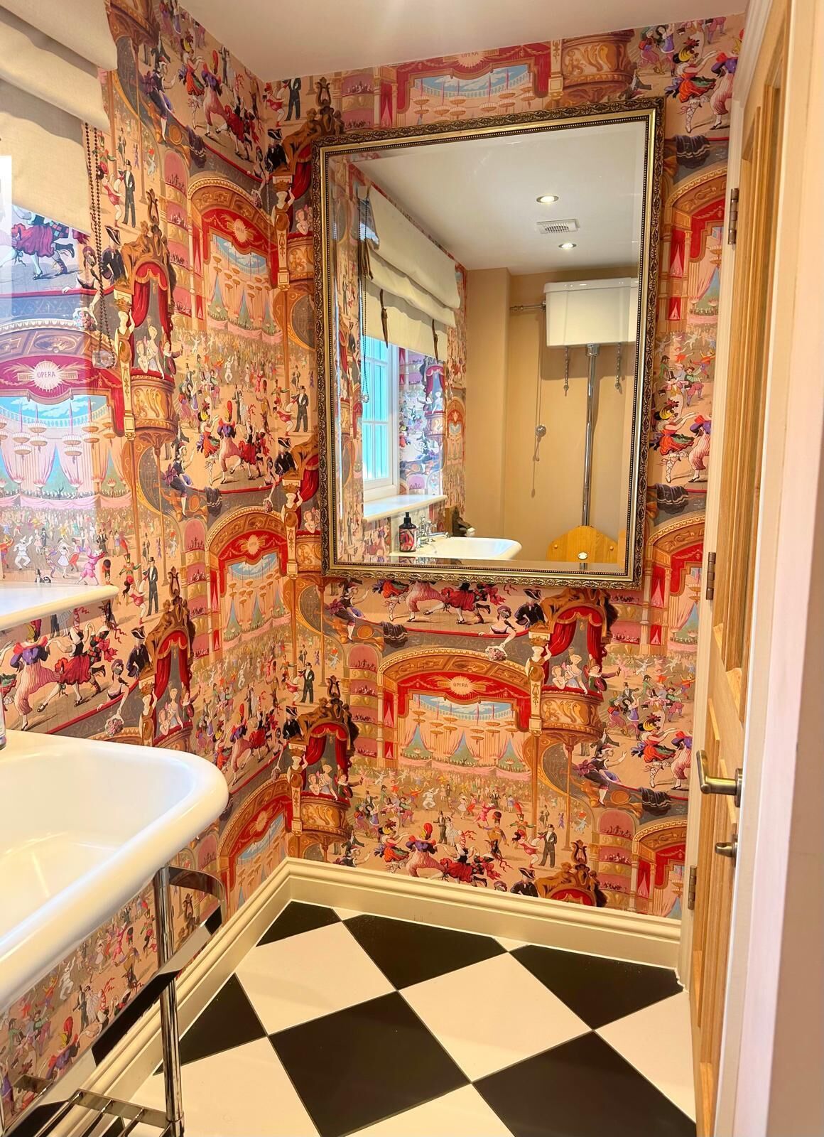 Ein Badezimmer mit Schachbrettmuster-Boden, einem Waschbecken und einem Spiegel. Die Wände sind mit farbenfrohen, detailreichen Wandgemälden verziert.