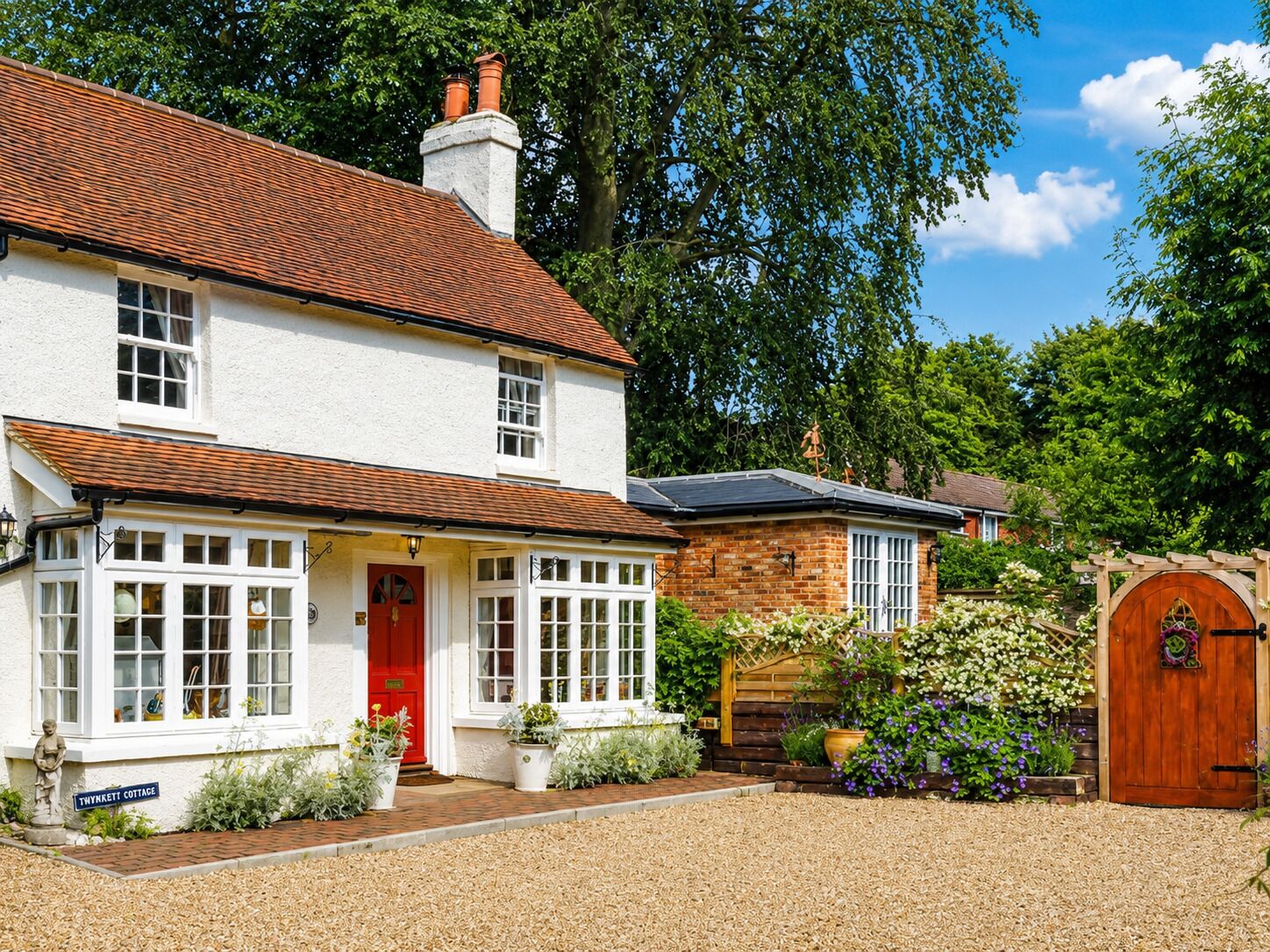 A two-story white house with a red door, a brick extension, and a garden with flowers and a wooden gate.