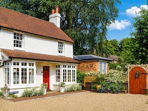 A two-story white house with a red door, a brick extension, and a garden with flowers and a wooden gate.