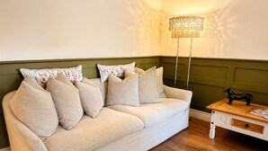 En beige soffa med flera kuddar, ett träbord med en tidning på och en golvlampa med en mönstrad lampskärm.