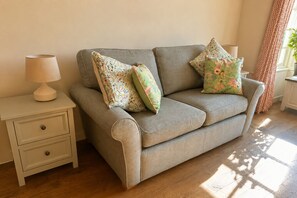 A neutral-toned sofa with floral-patterned cushions, a bedside table with a lamp, and a window with curtains.