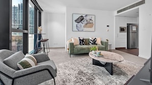 Un salon moderne avec un canapé vert, un fauteuil gris et une table basse. Une grande fenêtre offre une vue sur les bâtiments à l’extérieur.