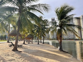 Uma praia com palmeiras, um pavilhão de colmo e um corpo d'água calmo.