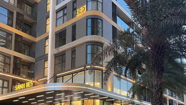 A modern hotel with a glass facade, a well-lit entrance, and a water feature in the foreground. - Vinhomes Grand Park-The Beverly Hills (Ho Chi Minh City)
