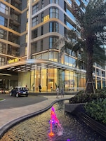 Um hotel moderno com fachada de vidro, entrada bem iluminada e um elemento de água em primeiro plano.
