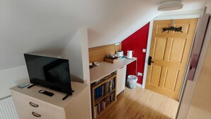 Een compacte kamer met een televisie op een dressoir, een boekenkast, een kitchenette met waterkoker en een deur naar een andere kamer.