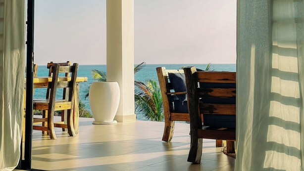 An open doorway leading to a balcony with wooden chairs and a white planter, overlooking a beach. - Villa Kweli (Makunduchi)