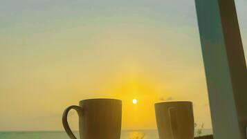 Dos tazas blancas sobre platillos en una mesa de madera, con vista a la puesta de sol sobre el océano.